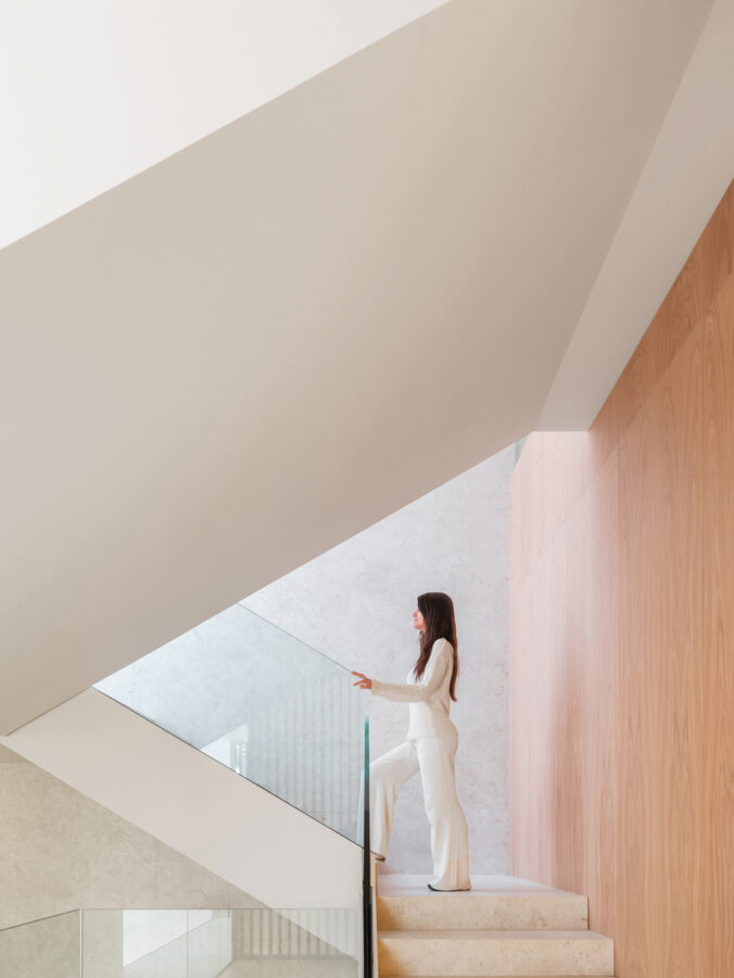 Escalera de porcelánico con barandilla de vidrio entre muros blancos y madera clara.